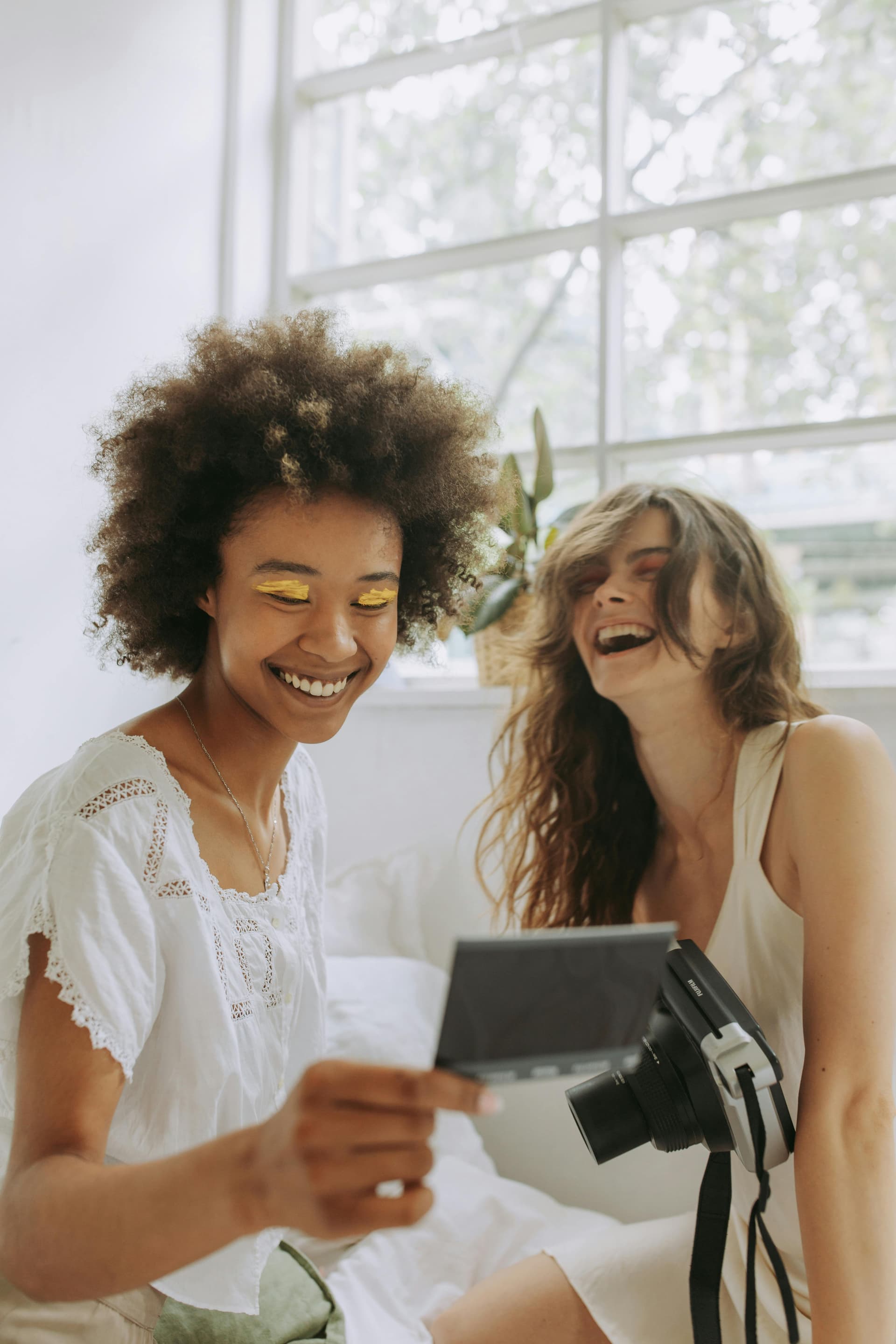 Two friends laughing while looking at a fresh instant photo in a bright bedroom.